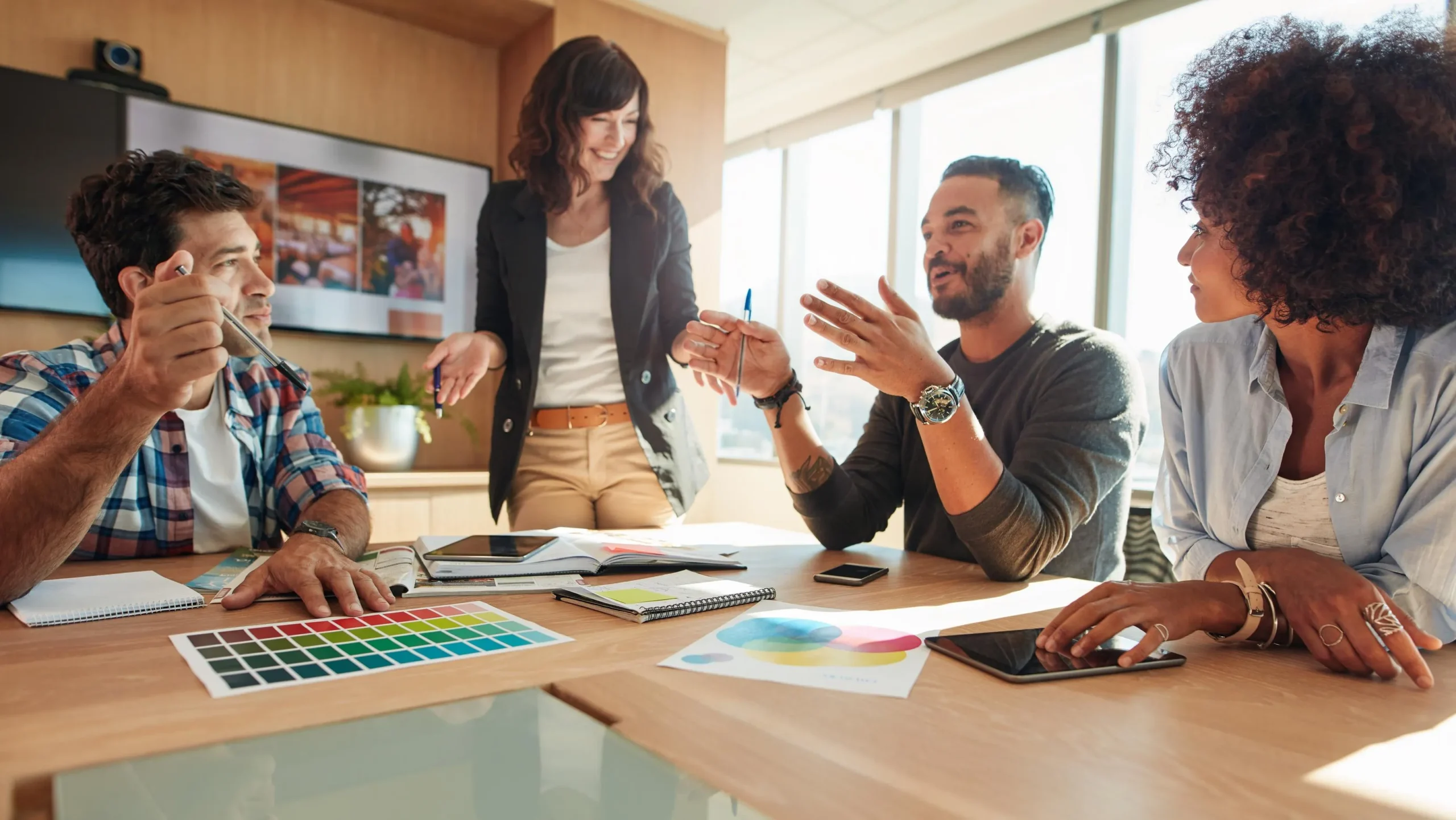 Group of four colleagues in a bright office discussing a project, with color swatches and papers on the table.