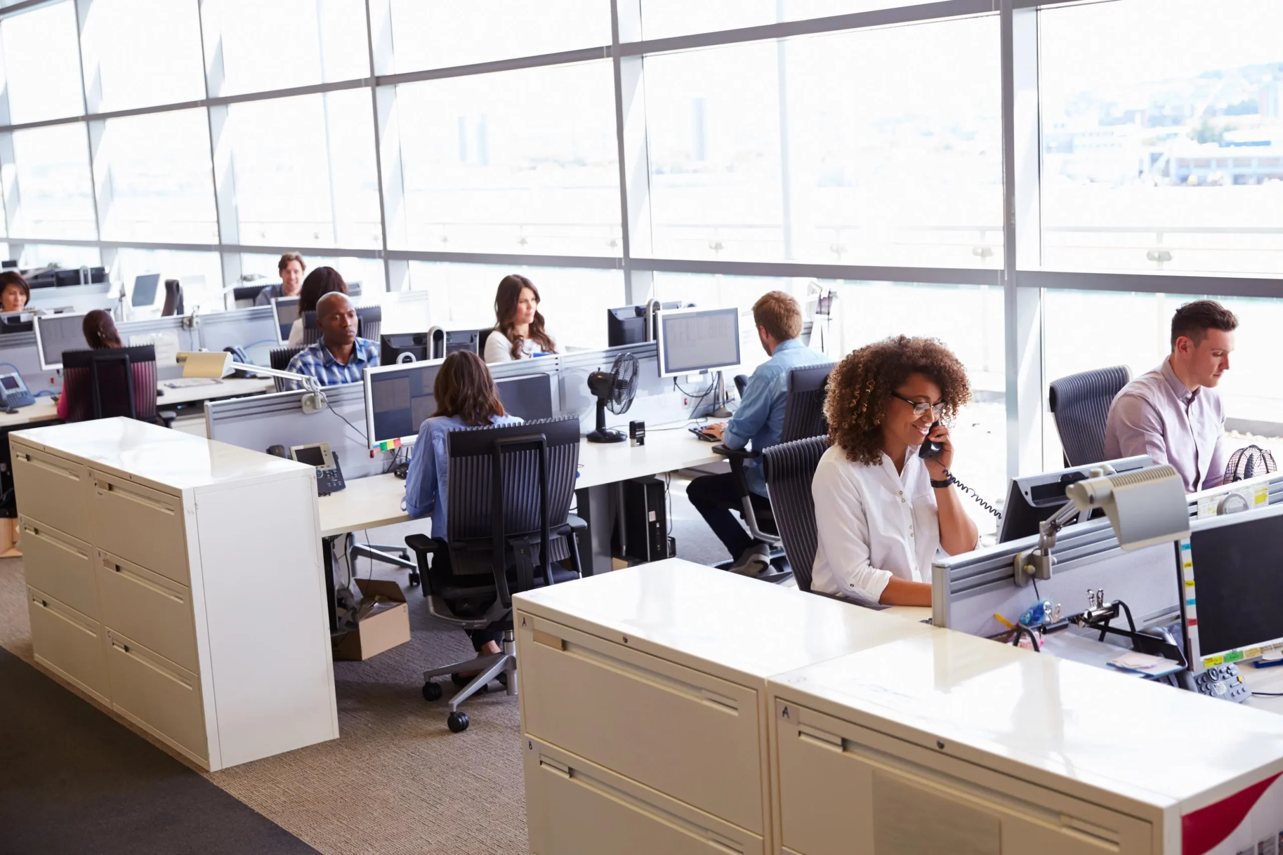 Open-plan office with many desks, computers, and employees working at stations by large glass windows in daylight.