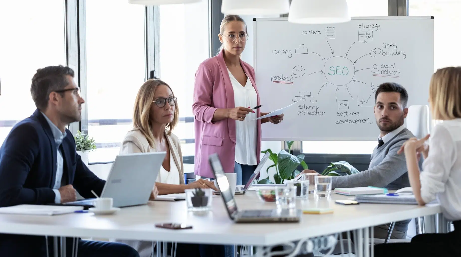 Woman in a pink blazer presenting SEO strategy to colleagues around a conference table.