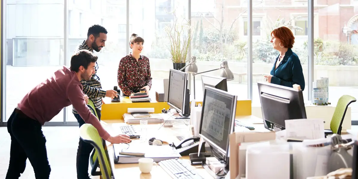 Five coworkers collaborate around a bright open-plan desk, discussing a project near computer monitors by a large windowed wall.