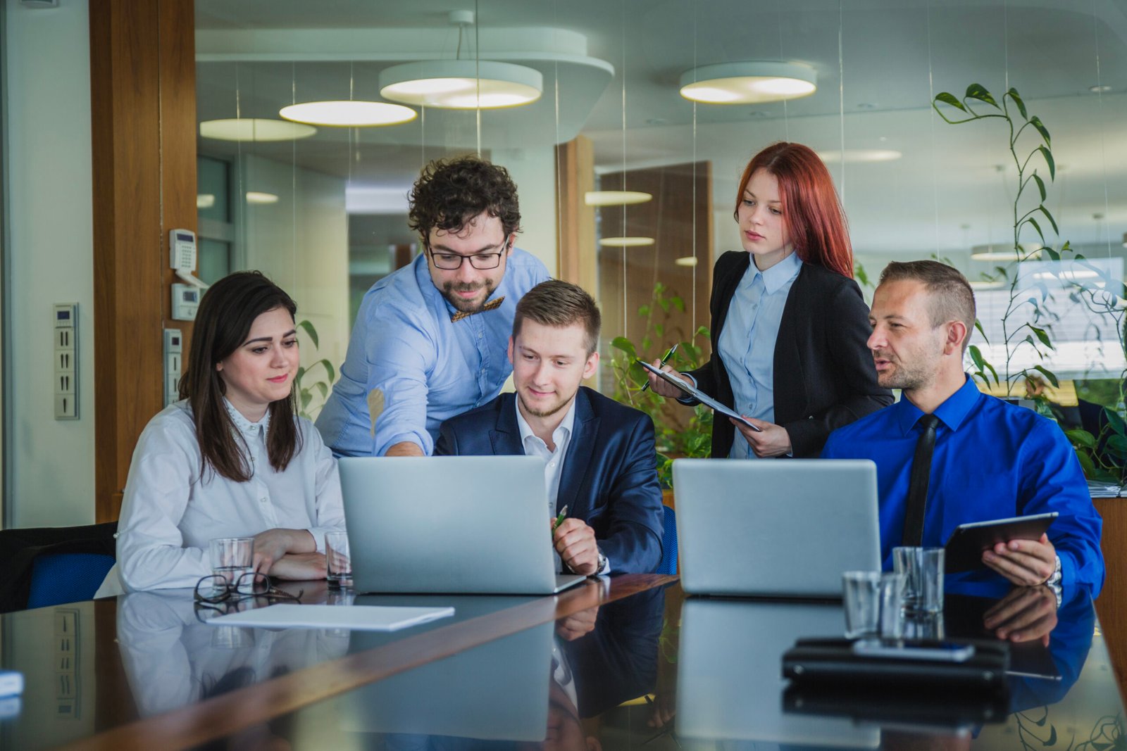 Five professionals collaborate around a glass conference table with laptops in a bright office space, reviewing documents.