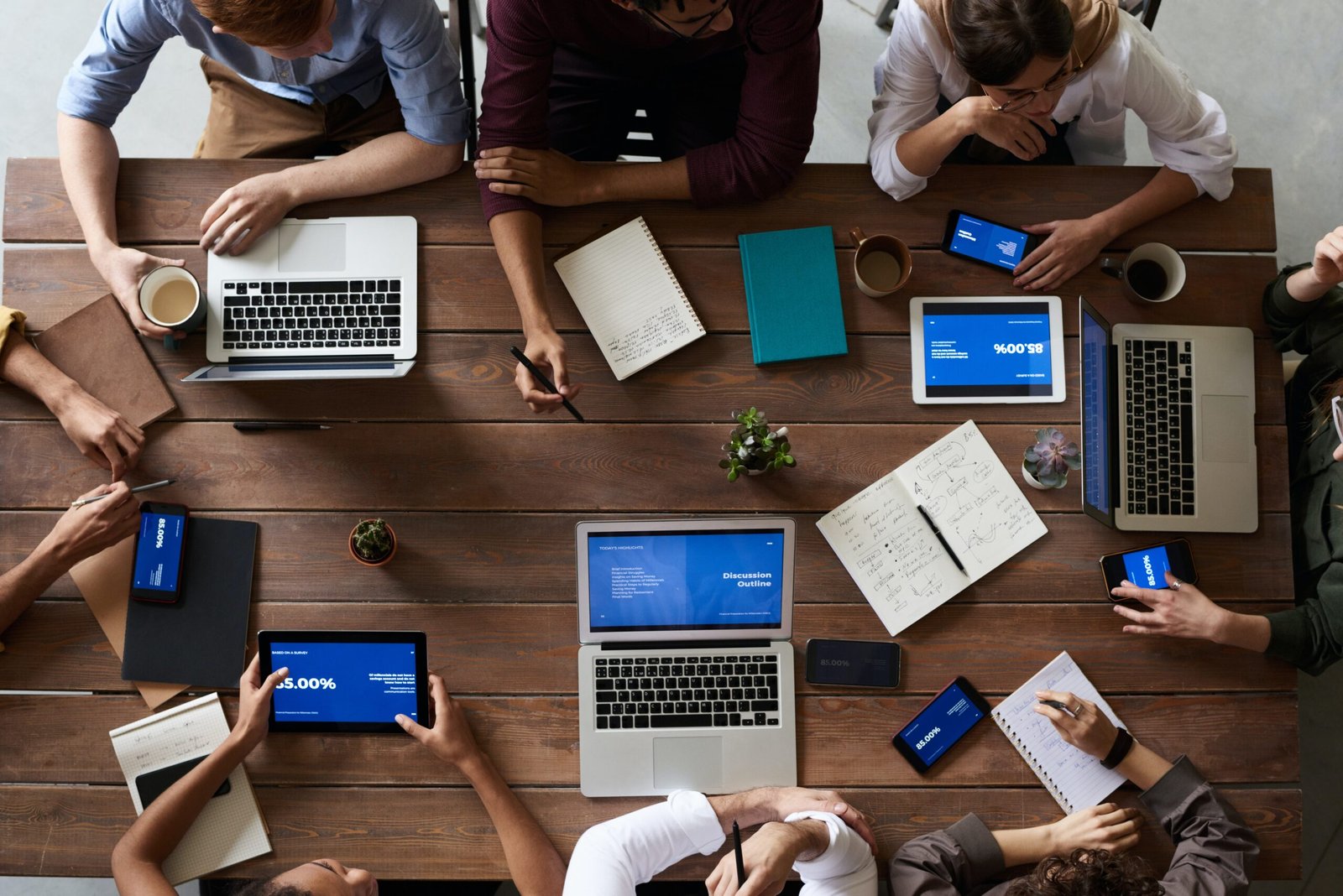 Overhead view of a collaborative meeting: people around a wooden table with laptops, tablets, notebooks, and coffee cups.