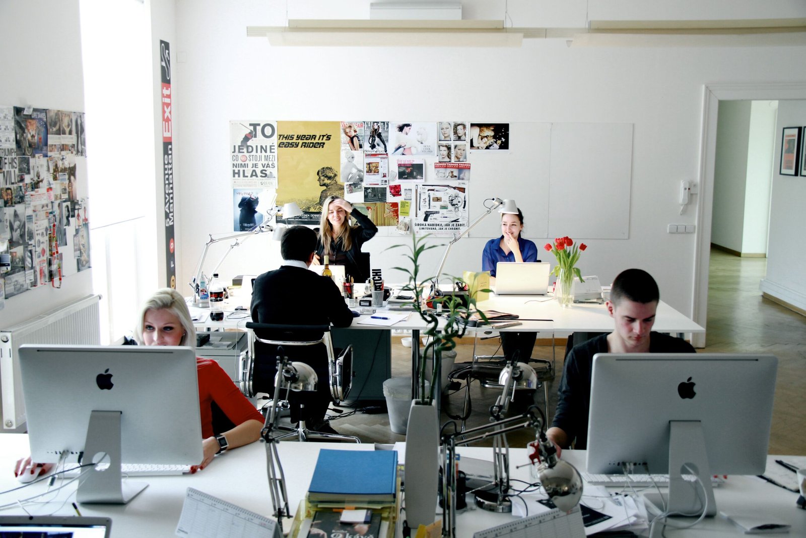 Open-plan office with several people working at desks and iMac computers, collage wall in the background and plants on desks in a bright room