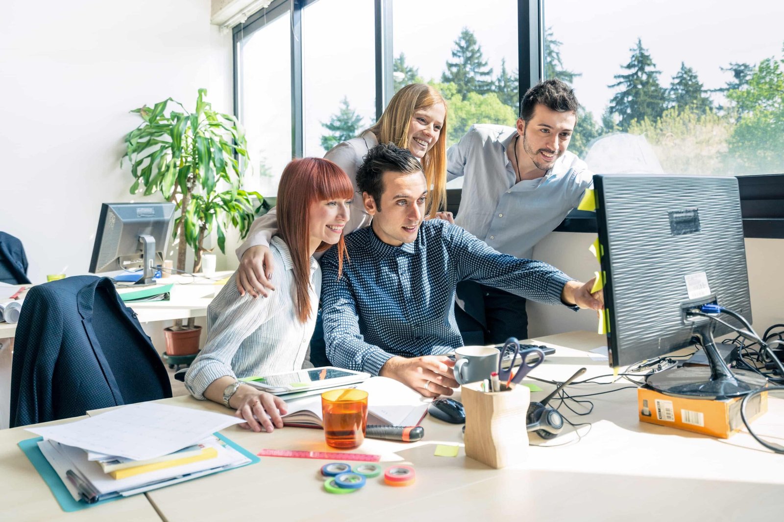 Four colleagues gather around a desk, looking at a computer monitor with smiles in a bright office. Papers, notebooks, and office supplies litter the desk, with a plant in the background and a large window behind them.