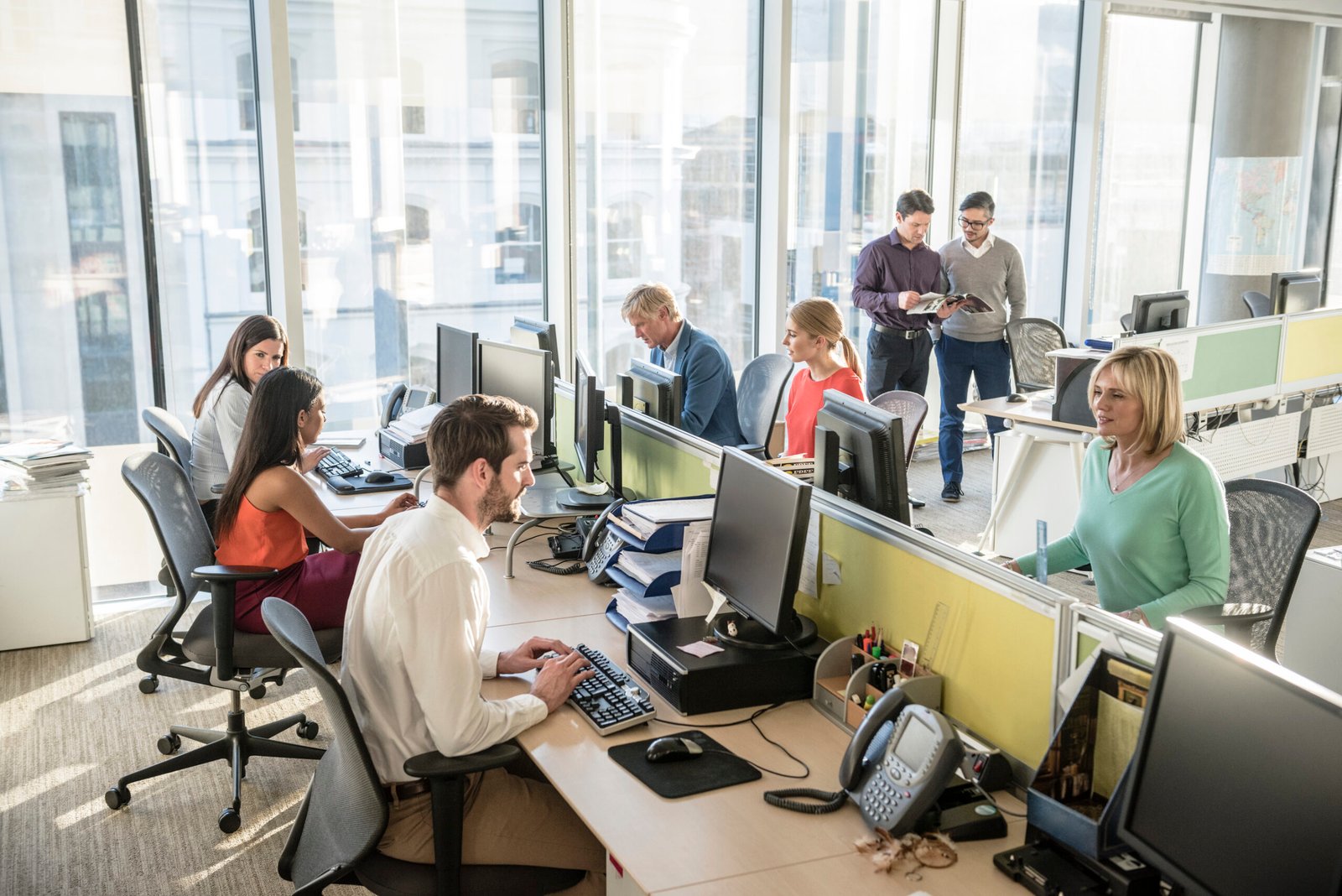 Open-plan office with workers at desks along a row of computers and two men standing and looking at a magazine by the windows, bright natural light.