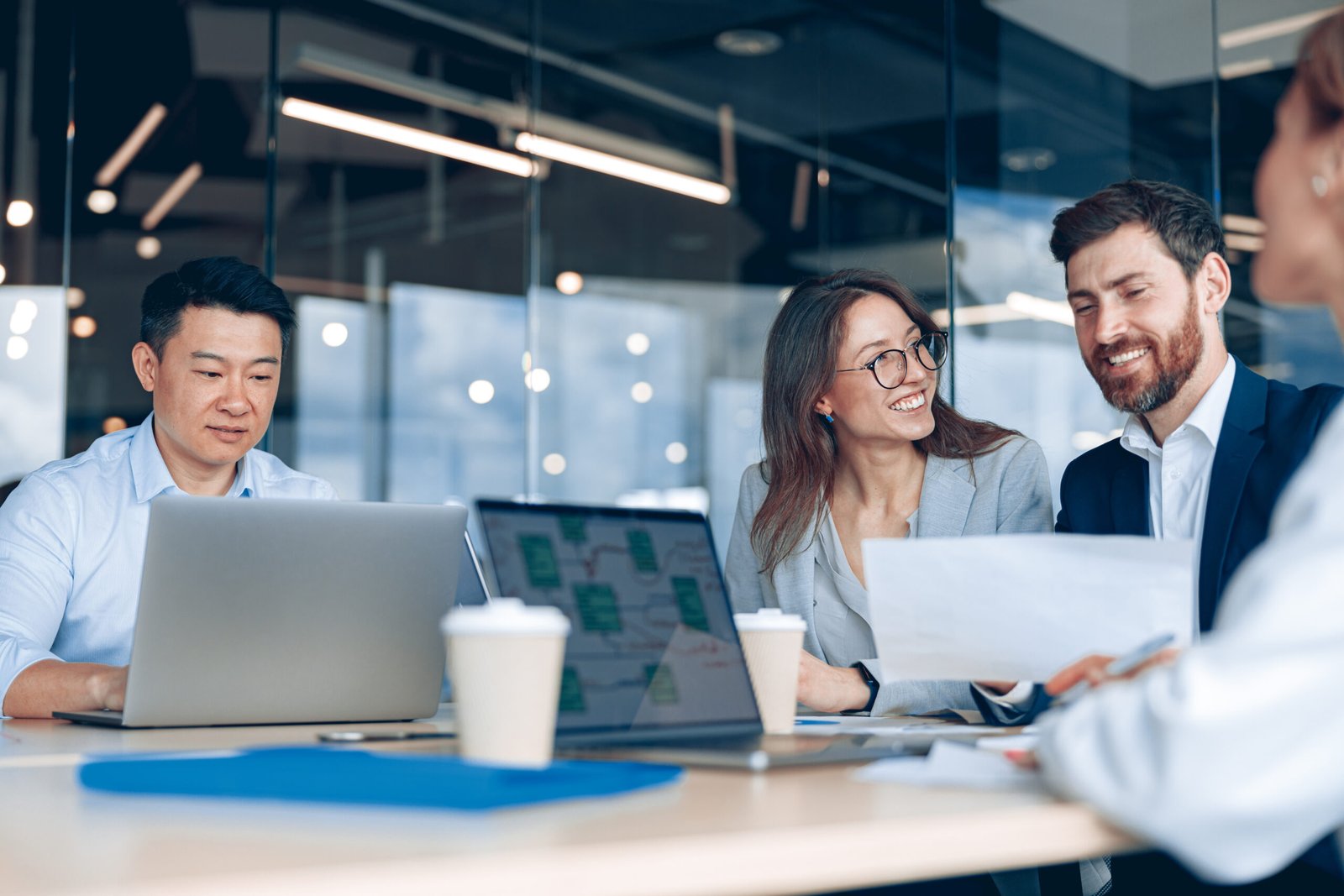 Group of diverse coworkers collaborating around a table with laptops and coffee cups in a modern office setting