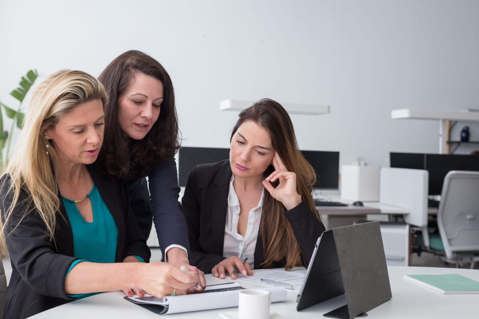 Three women in business attire collaborate at a desk, reviewing papers and looking at a laptop in a bright office.