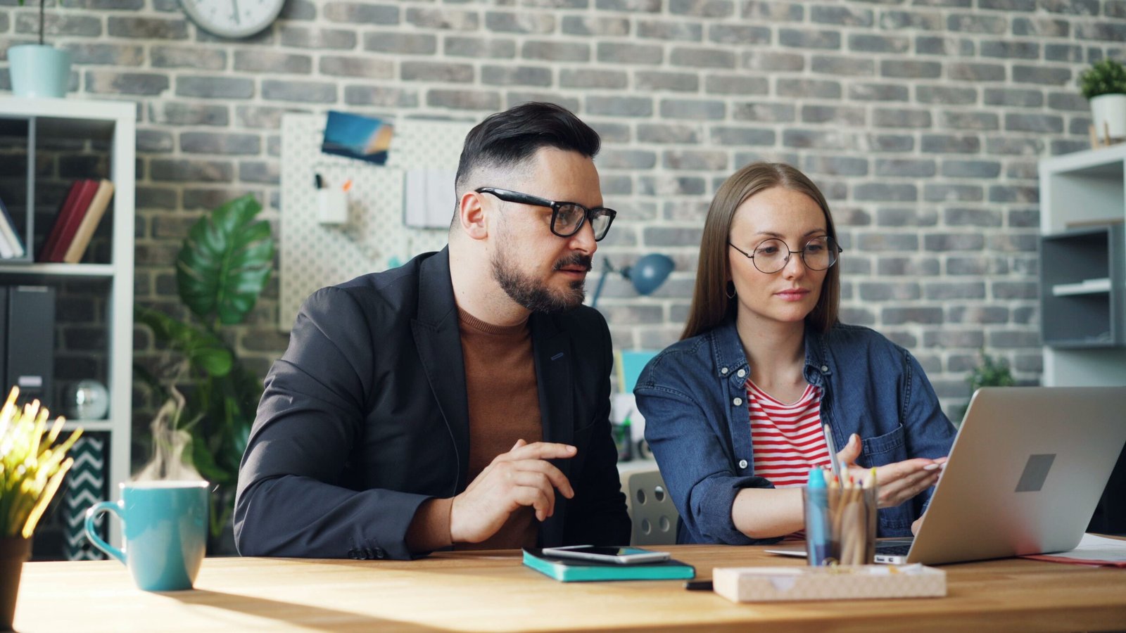 Two colleagues collaborate at a wooden desk with a laptop in a modern office setting.