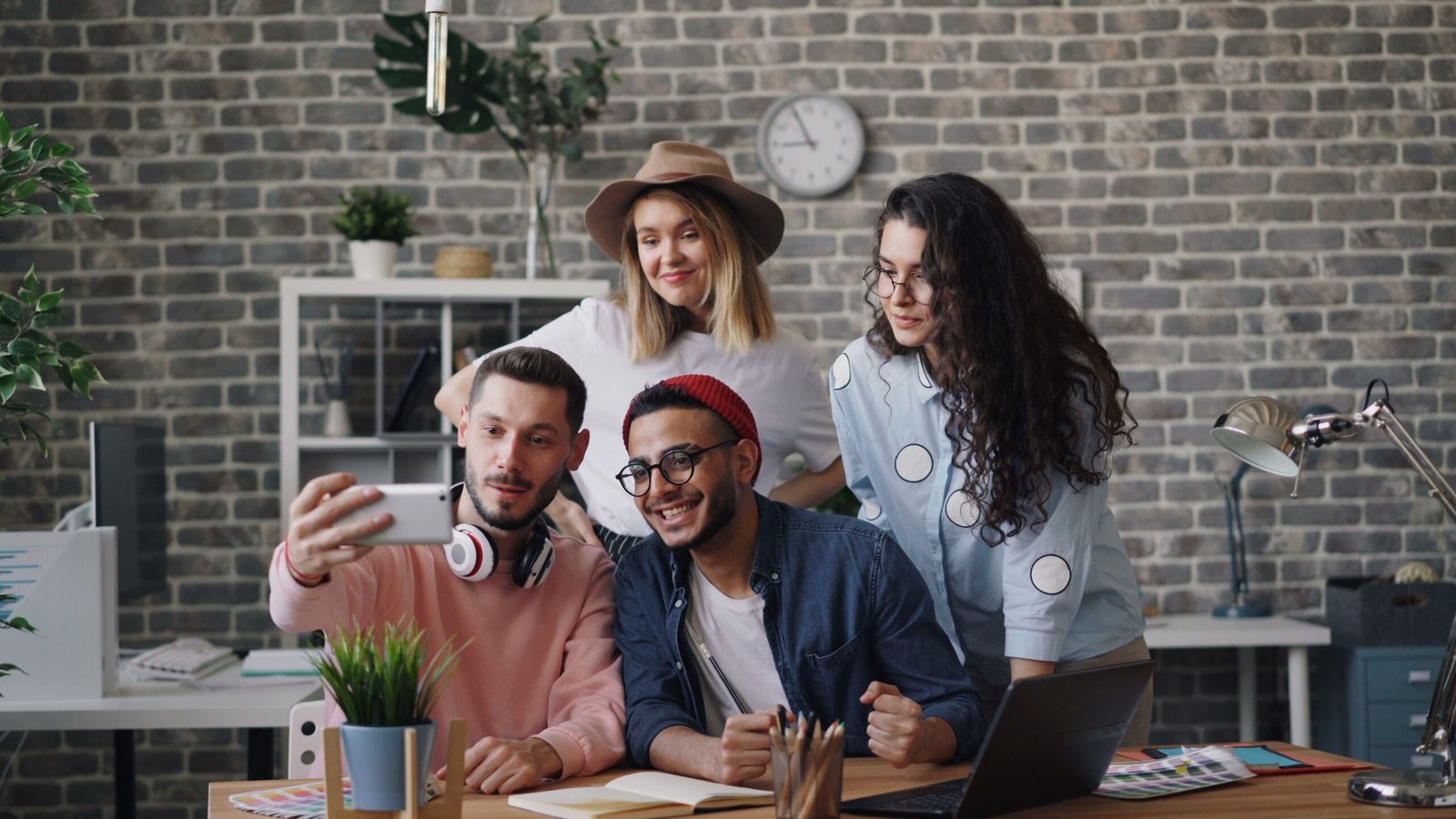 Group of four friends taking a selfie in a modern office with brick wall, plants, and a laptop on the desk behind them.”