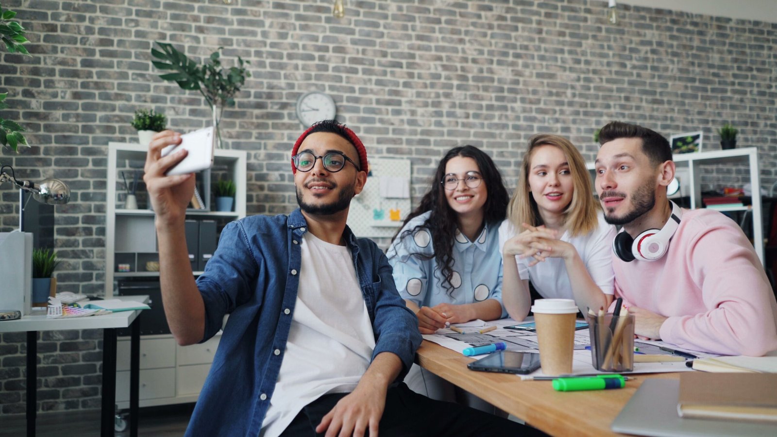 Four friends or coworkers take a group selfie at a cluttered office desk using a smartphone.