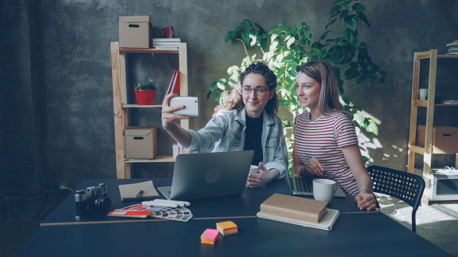 Two women sit at a desk in a modern office, taking a selfie with a smartphone while a laptop and notebooks are nearby.