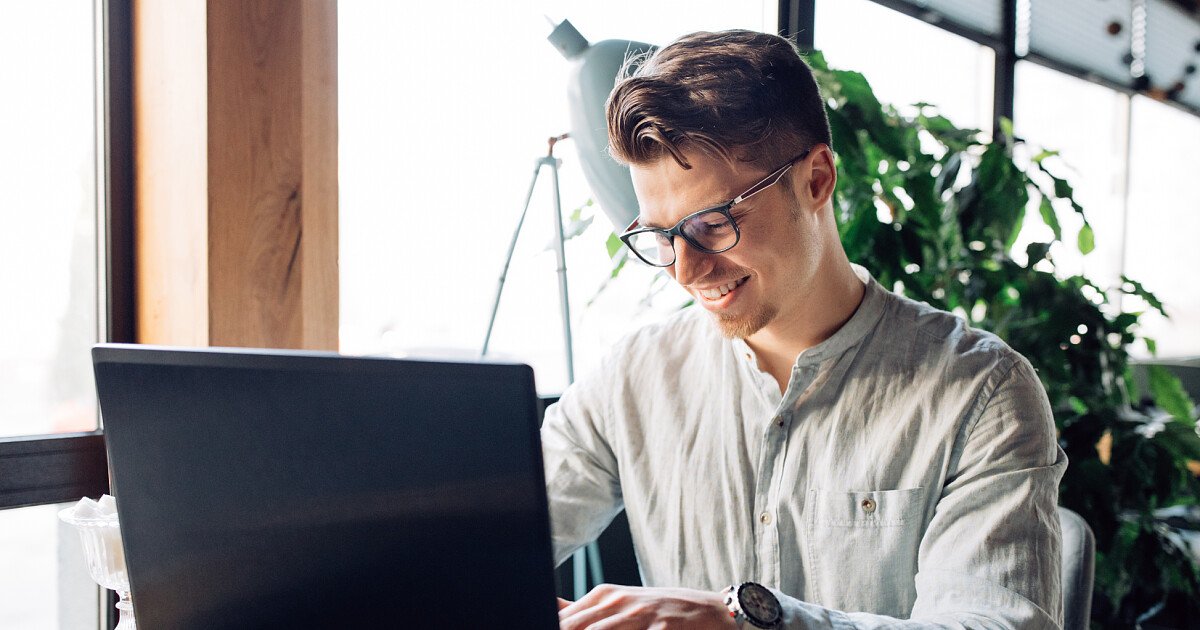 Man with glasses typing on a laptop at a bright desk with a plant in the background while smiling.