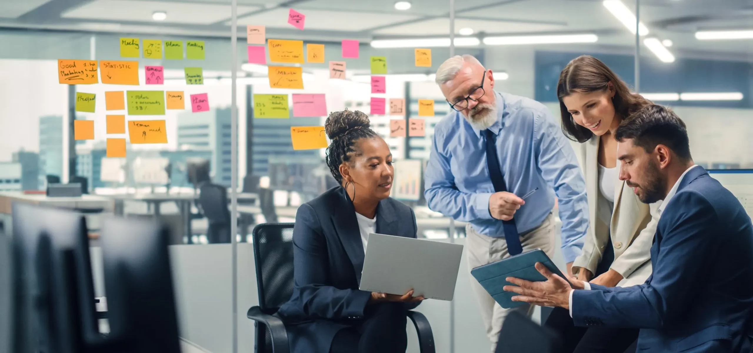 Team of four professionals collaborating around a desk, reviewing notes on a glass wall covered with colorful sticky notes.