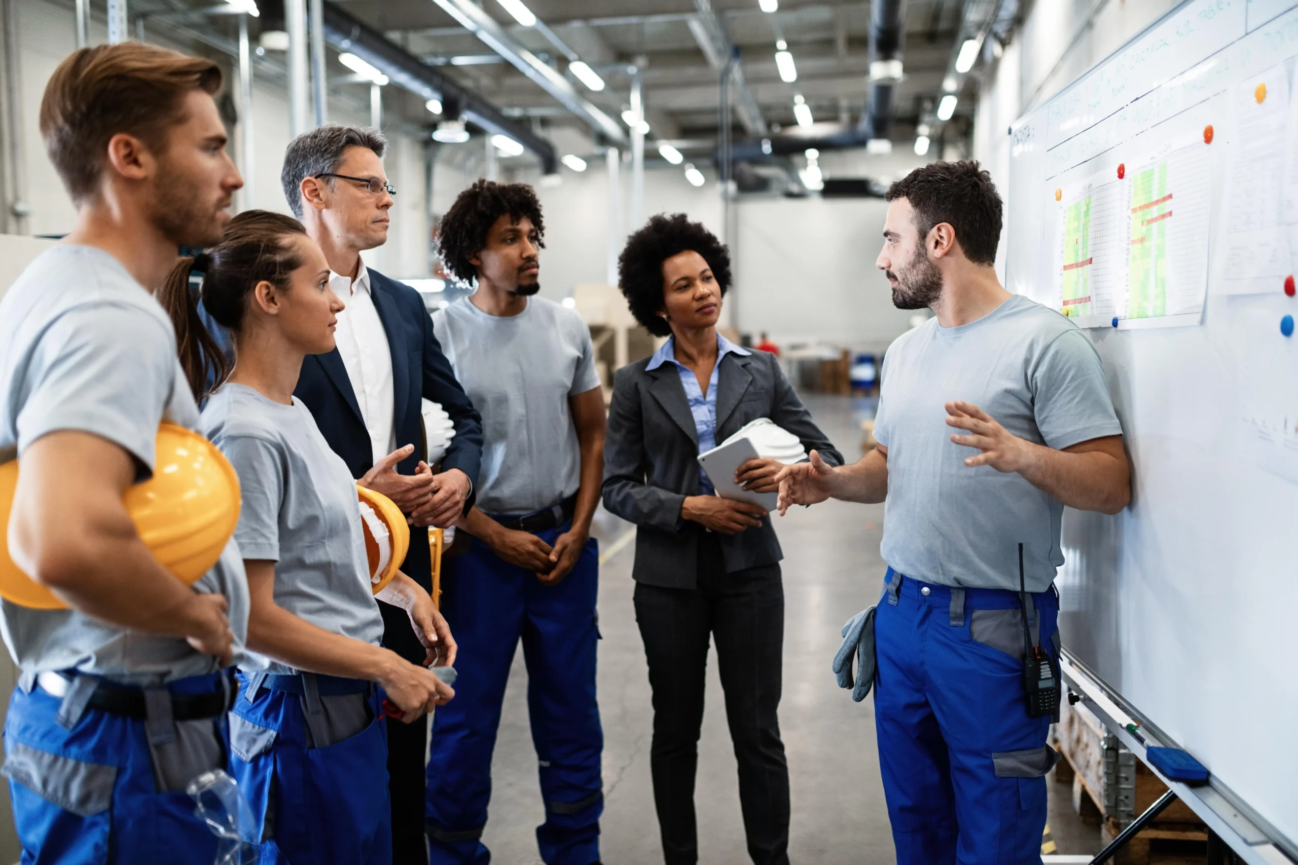 Group of diverse factory workers listening to a presenter by a whiteboard with charts in an industrial setting.