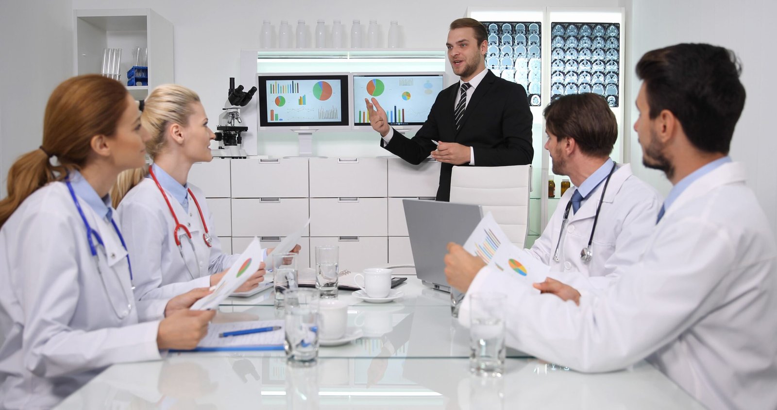 Medical team in white coats sits around a glass conference table while a presenter explains charts on screens at the front of the room, with brain scans on a display to the side.