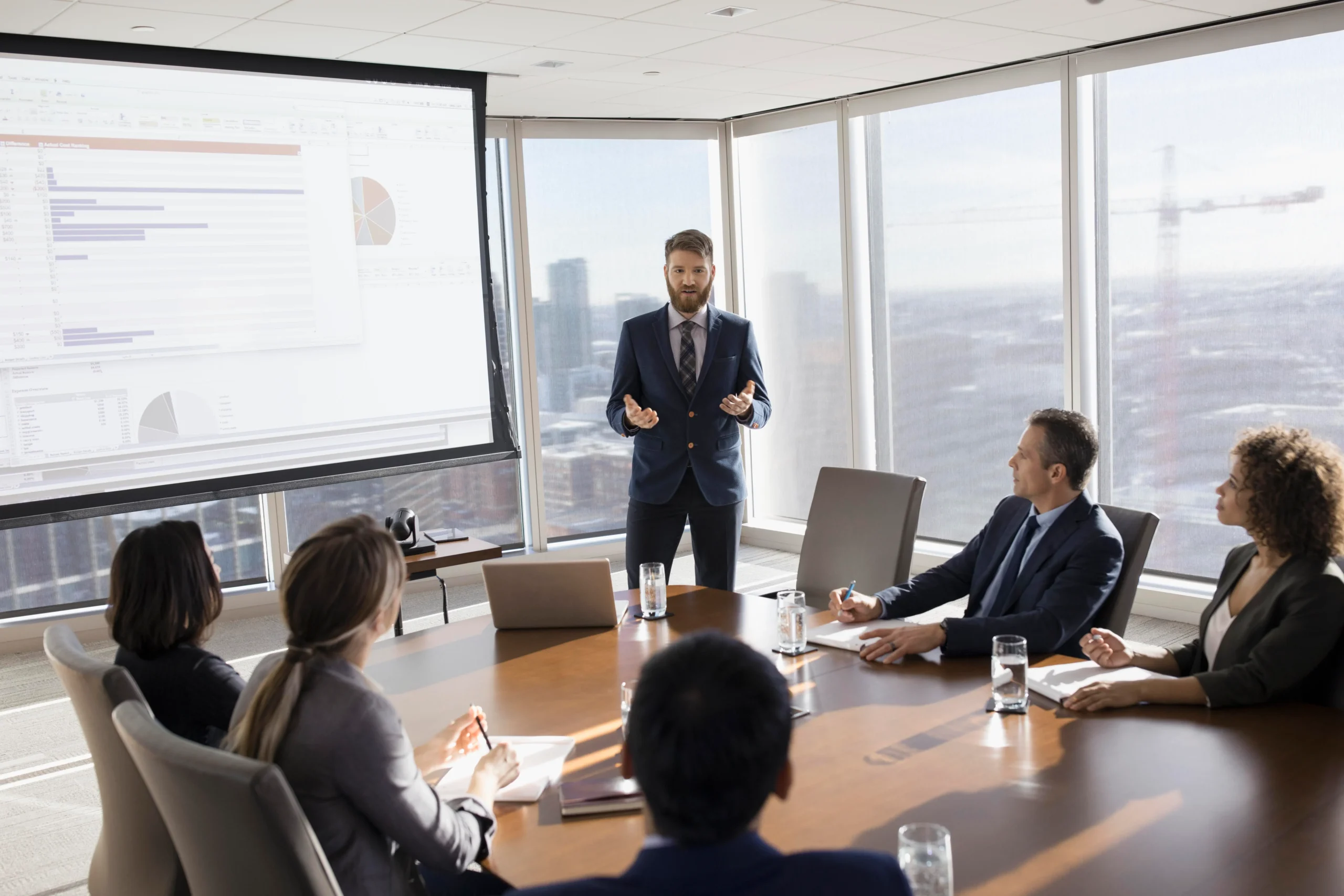 Business presenter in a navy suit explains charts to colleagues around a large conference table with a projector screen nearby.