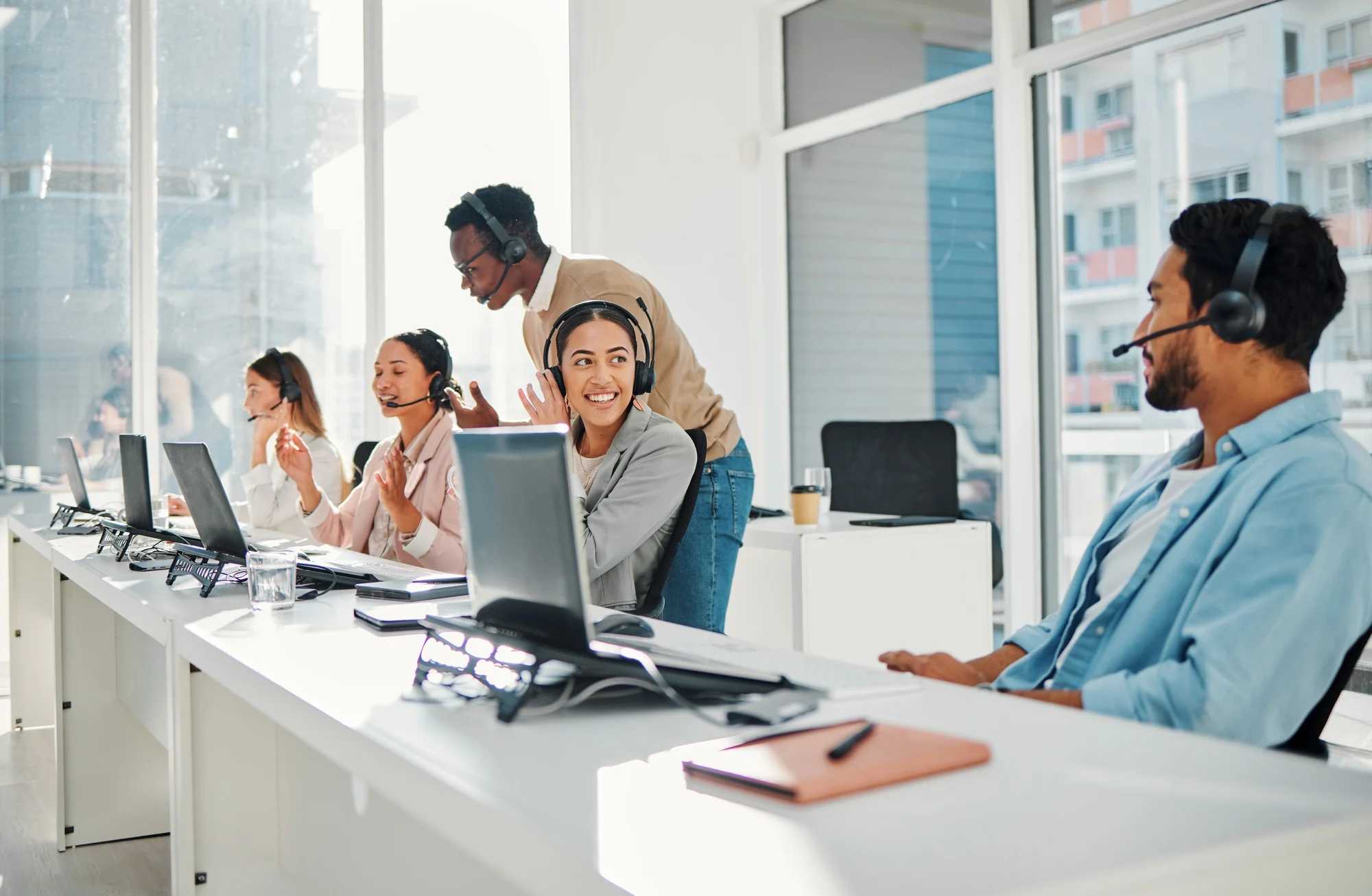 Smiling woman wearing a headset at a bright call center desk with colleagues in the background.