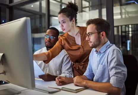 Three colleagues collaborate at a large desktop computer in a modern office; a woman points at the screen while two men look on nearby.