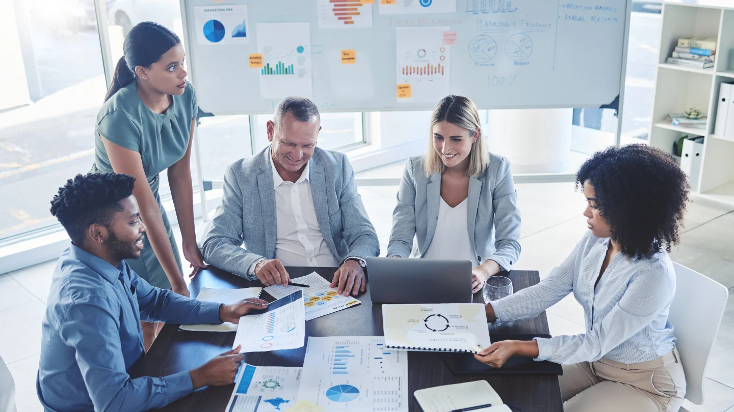Five diverse professionals collaborate around a table with charts, graphs, and a laptop in a bright office setting.