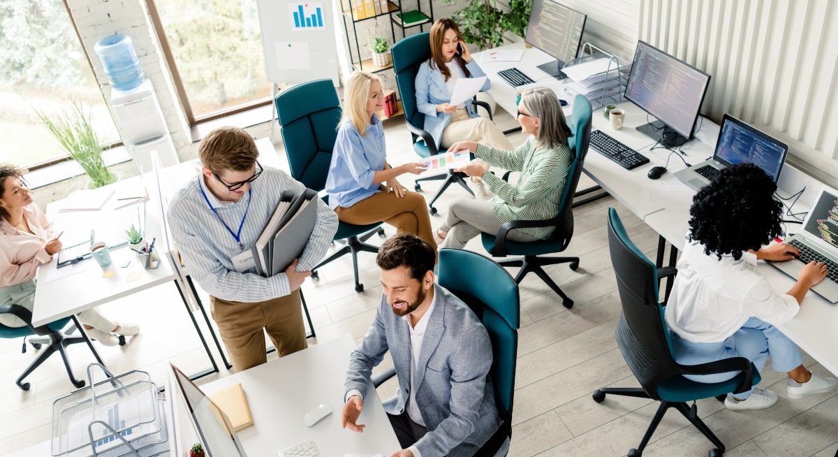 A busy open-plan office with several coworkers collaborating: people sit at desks with computers, some discussing charts and documents in a bright, modern space.