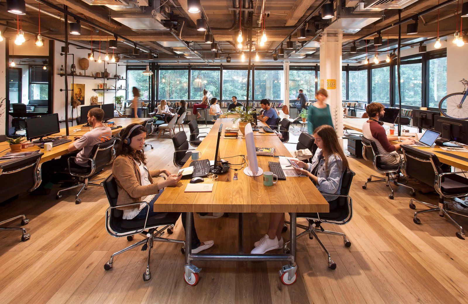 Open-plan coworking office with people working at long wooden tables, laptops, warm hanging bulbs, and large windows