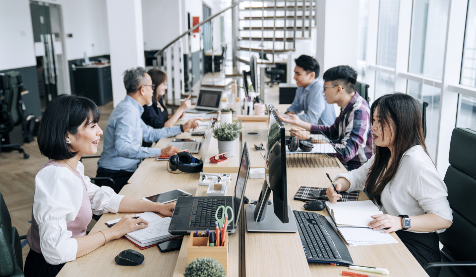 Team of coworkers seated along a long desk in a bright open-plan office, working on laptops and taking notes.
