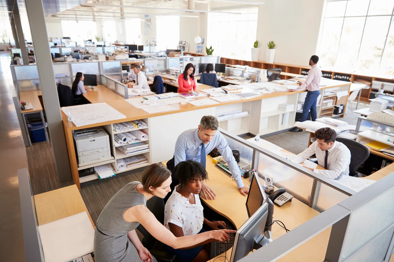 Team of colleagues huddle around a computer at a shared desk in a bright open-office.