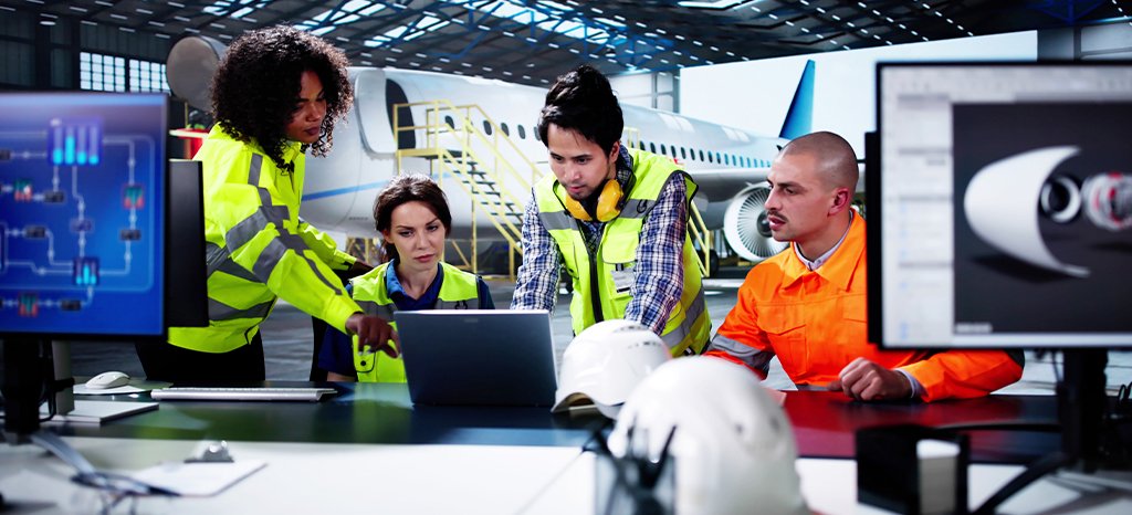 Aviation maintenance team in high-visibility vests huddle around laptops and monitors in a hangar with a jet in the background.