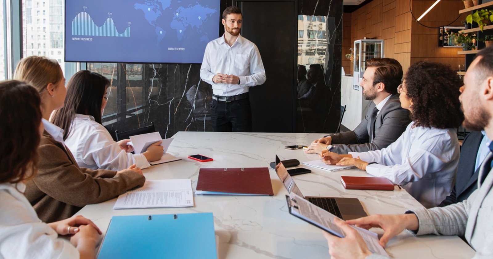 Man standing and presenting to a seated group around a marble conference table, with a world map/chart on a screen behind him
