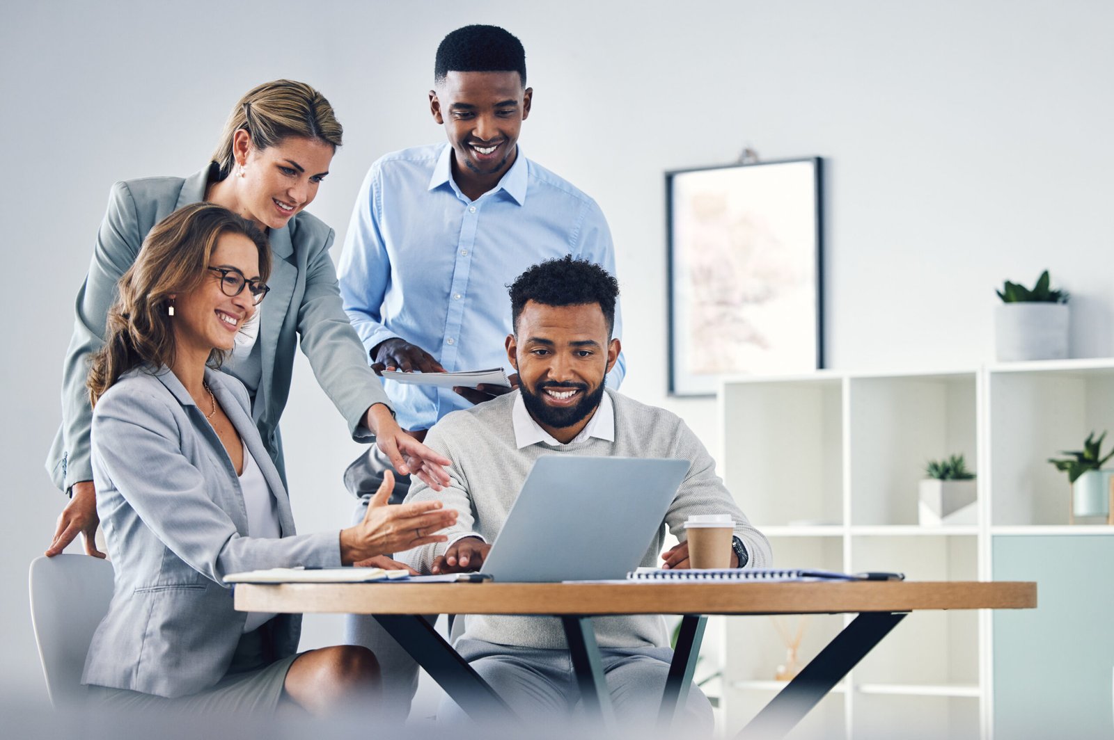 Four coworkers of diverse ages lean over a desk, reviewing a laptop and discussing.