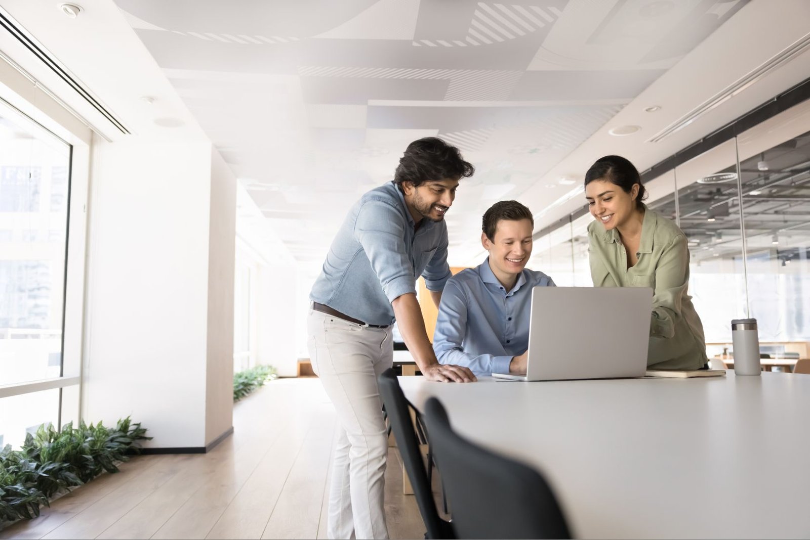 Three coworkers gather around a laptop at a long conference table, smiling as they review something together in a bright office.