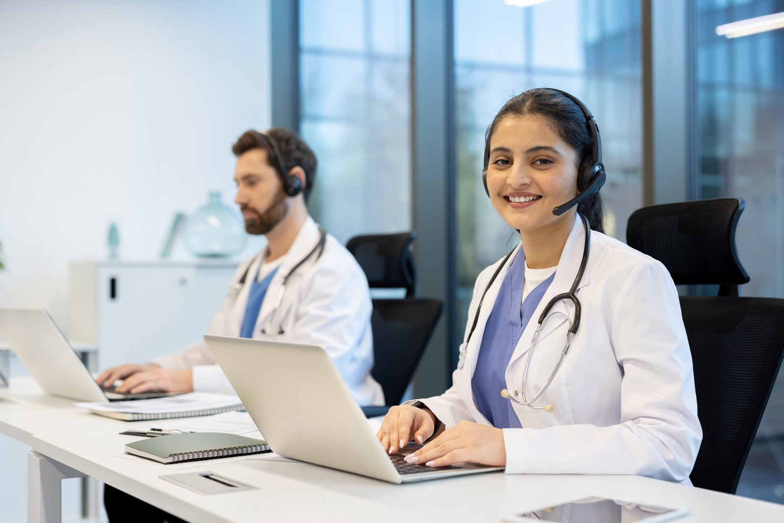 Two healthcare workers in white coats at a desk with laptops and headsets, smiling at the camera in a bright call-center setting (customer support/triage).