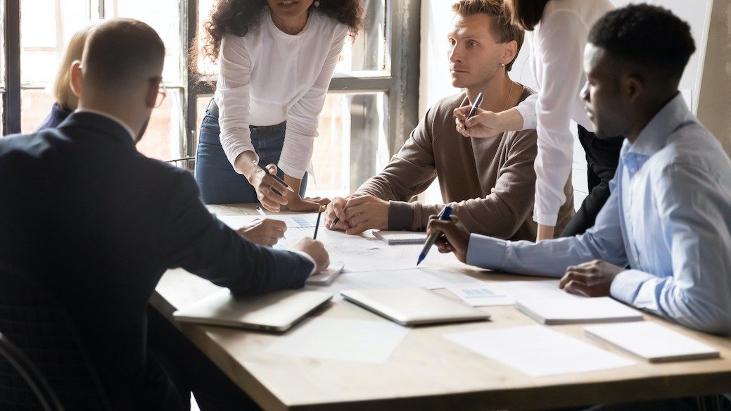 Six people collaborate around a table with papers, notebooks, and pens in a bright office. (informative)