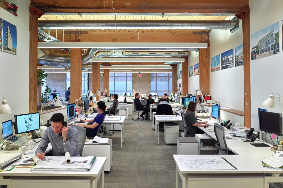 Open-plan design studio with engineers and designers at desks, blueprints and computers visible across the room.