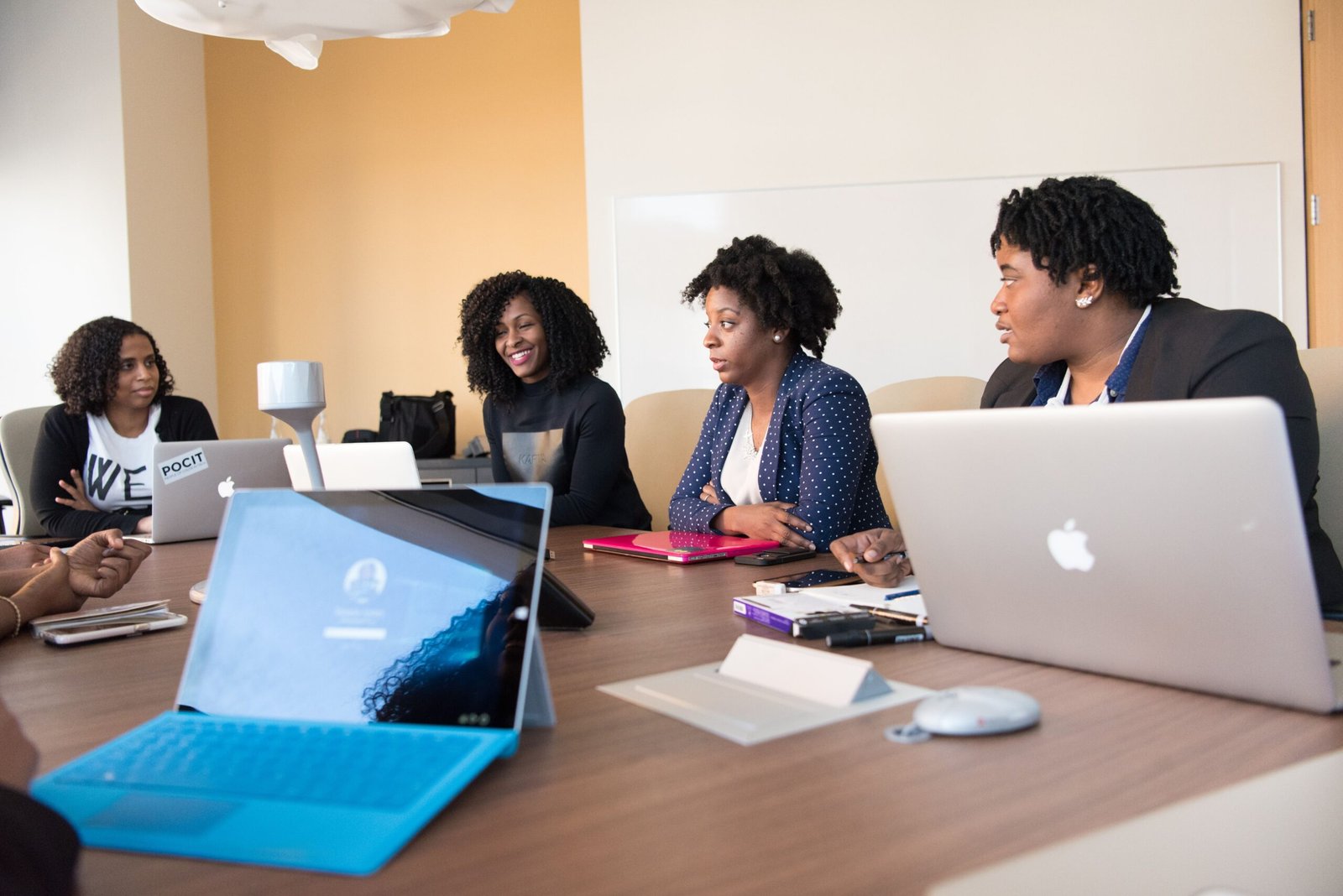 Four women sit around a conference table with laptops and notebooks, engaging in a collaborative meeting in a bright office.