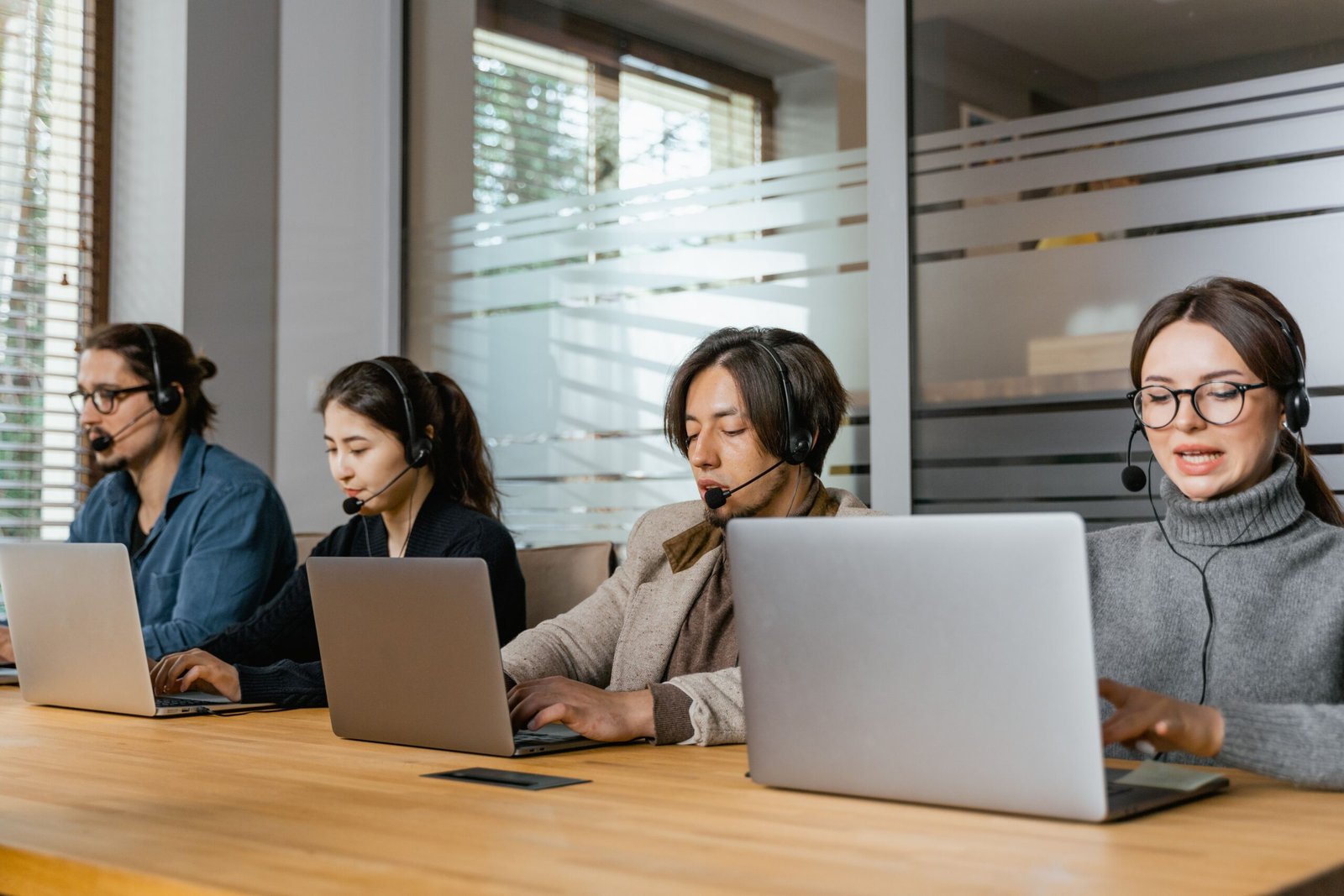 Four coworkers wearing headsets and using laptops at a long wooden table in an office.