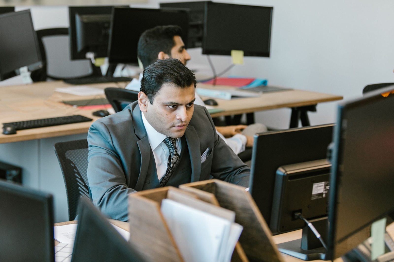 Man in a gray suit and tie works at a desk in an open-plan office with multiple computer monitors around him.