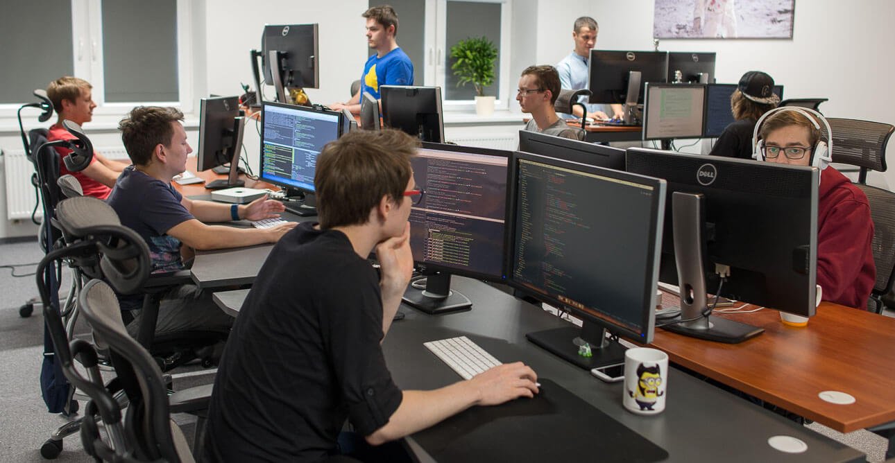Open-plan office with several people coding at desks, multiple monitors displaying code.