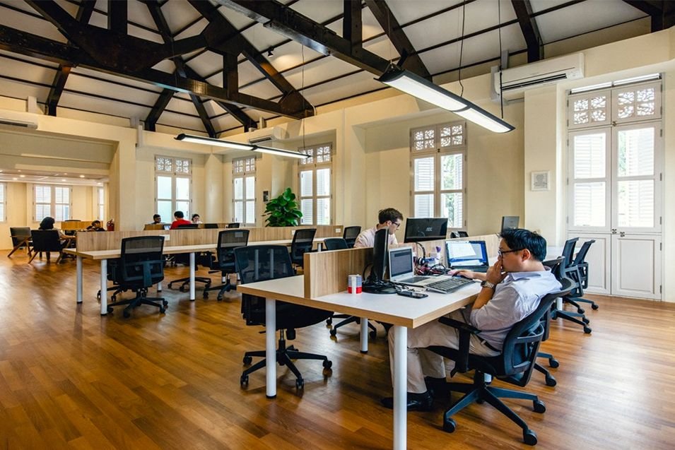 Open-plan coworking space with people working on laptops at desks; sunlit room with windows, wooden floor, and exposed beams.