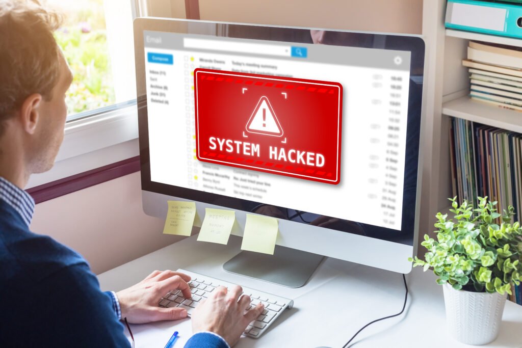 Person typing at a desk; monitor shows a red warning sign 'SYSTEM HACKED' on the screen.