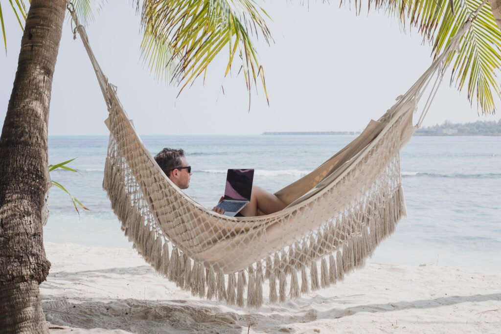 Man relaxing in a woven hammock between palm trees on a sandy beach, using a laptop and tablet at the water's edge.