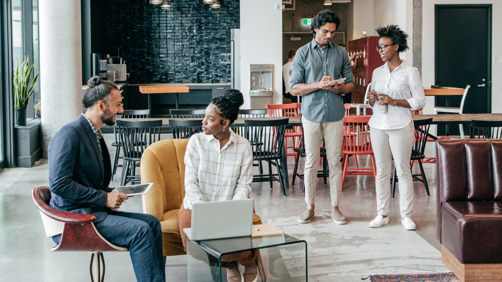 Four coworkers in a modern open-office lounge: a man in a blue blazer sits with a tablet, a woman with a laptop sits on a yellow chair, and two others stand nearby chatting, holding drinks.