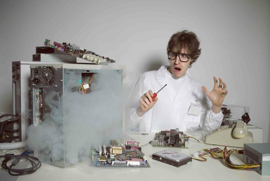 Scientist in safety goggles and white coat holds a screwdriver as a desktop computer hisses with smoke from an open case nearby.