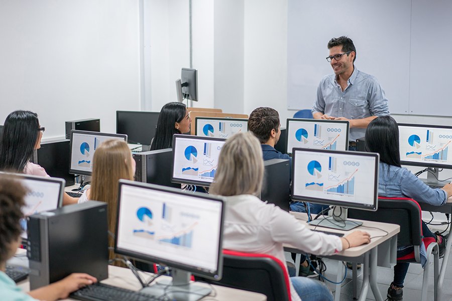 Instructor leads a data-analytics class in a computer lab, students focused on monitors displaying charts and graphs.