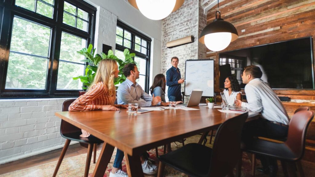 Diverse team of coworkers in a bright loft-style conference room around a wooden table, with a presenter at a whiteboard.