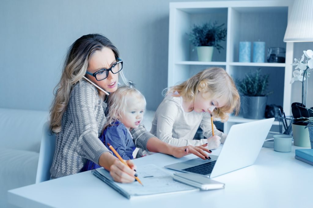Mother wearing glasses on a phone while two young children do homework at a white desk with a laptop and pencils nearby.