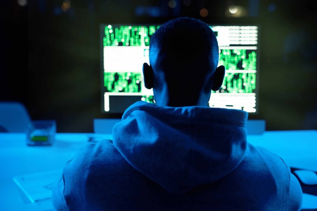 Person in a hoodie sits with back to the camera, looking at multiple monitors displaying neon green code in a dark room.
