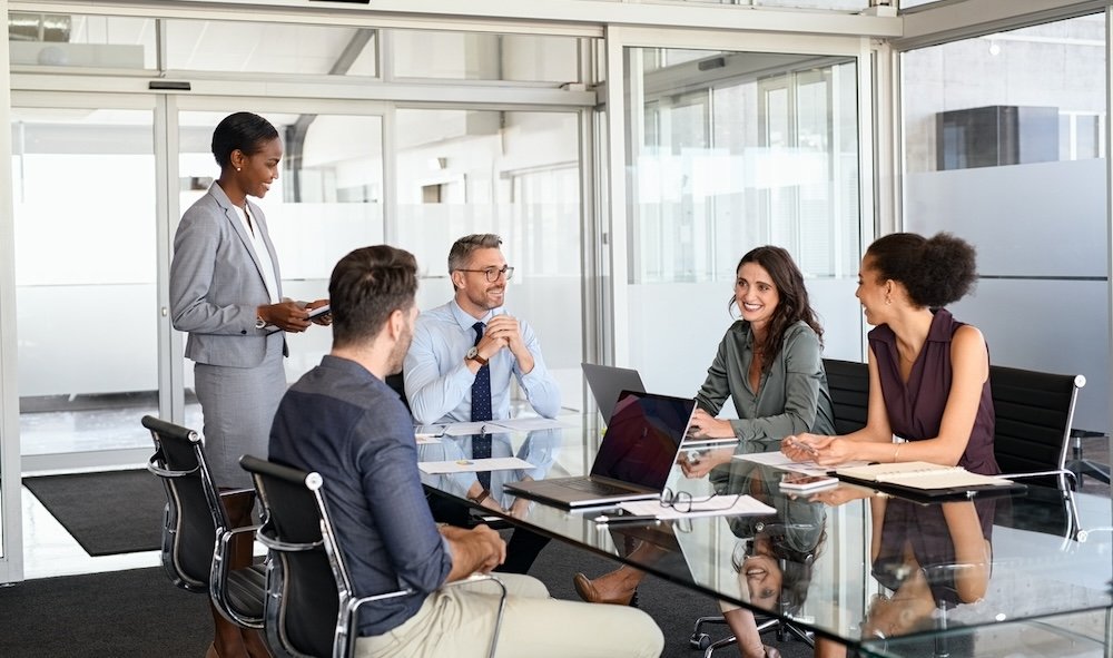 Diverse team in a glass-walled conference room around a glass table, one woman standing and presenting to colleagues with laptops open.