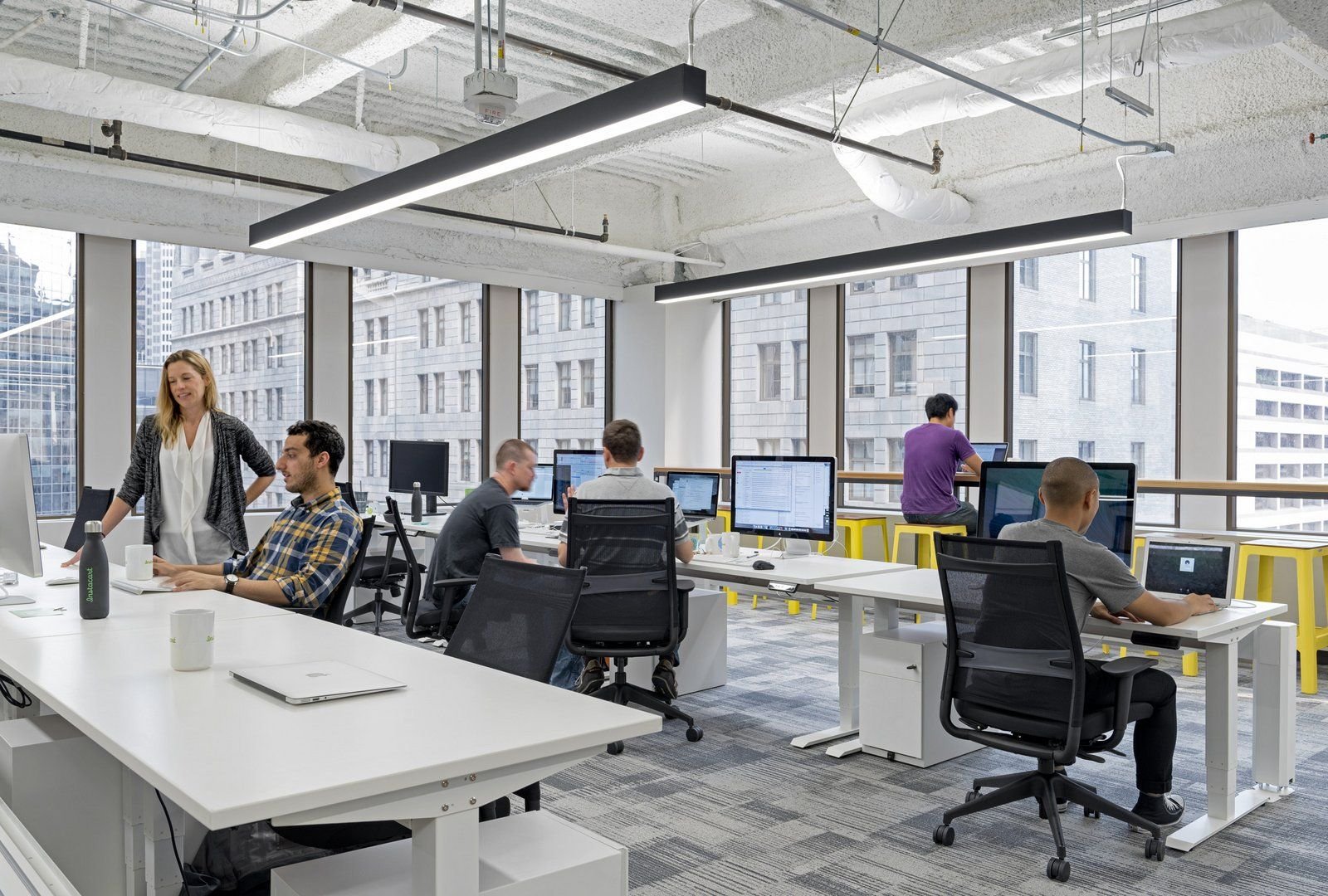 Open-plan office with several employees at white desks and large windows overlooking a cityscape, modern exposed ceiling.