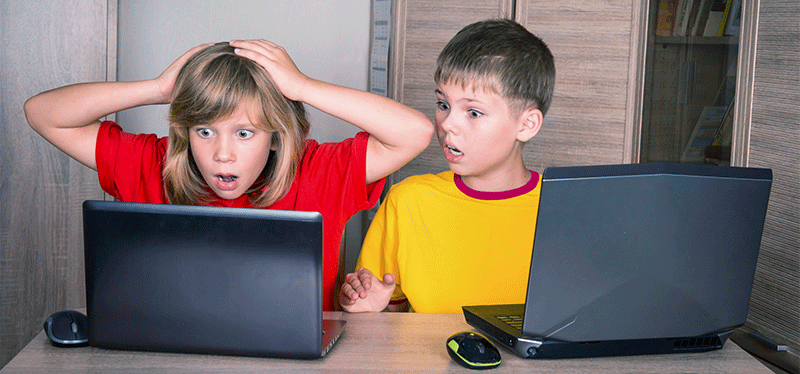 Two children sit at a desk with laptops, looking shocked at what’s on their screens.